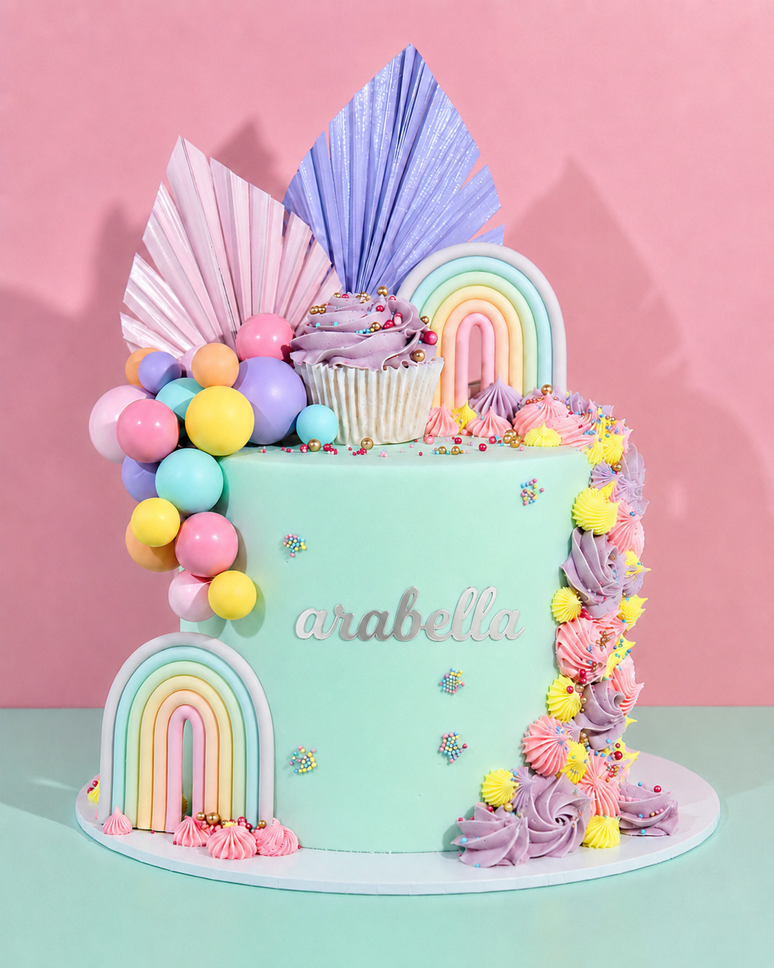A pastel-colored decorated cake on a matching round cake board, set against a pink background. The cake is mint green with silver 'arabella' lettering, decorated with rainbow arches, pastel balloons, a cupcake topper, and piped frosting swirls in pink, purple, and yellow.