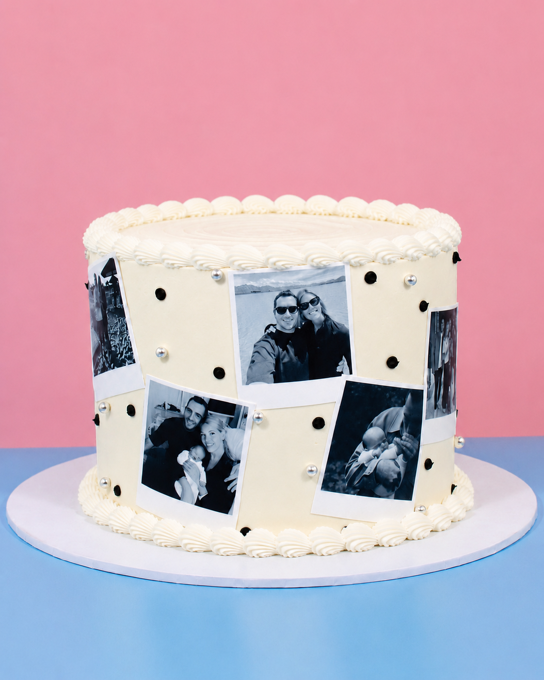A round white frosted cake decorated with black-and-white photo prints and small silver dots, displayed on a white round board against a pink and blue background.