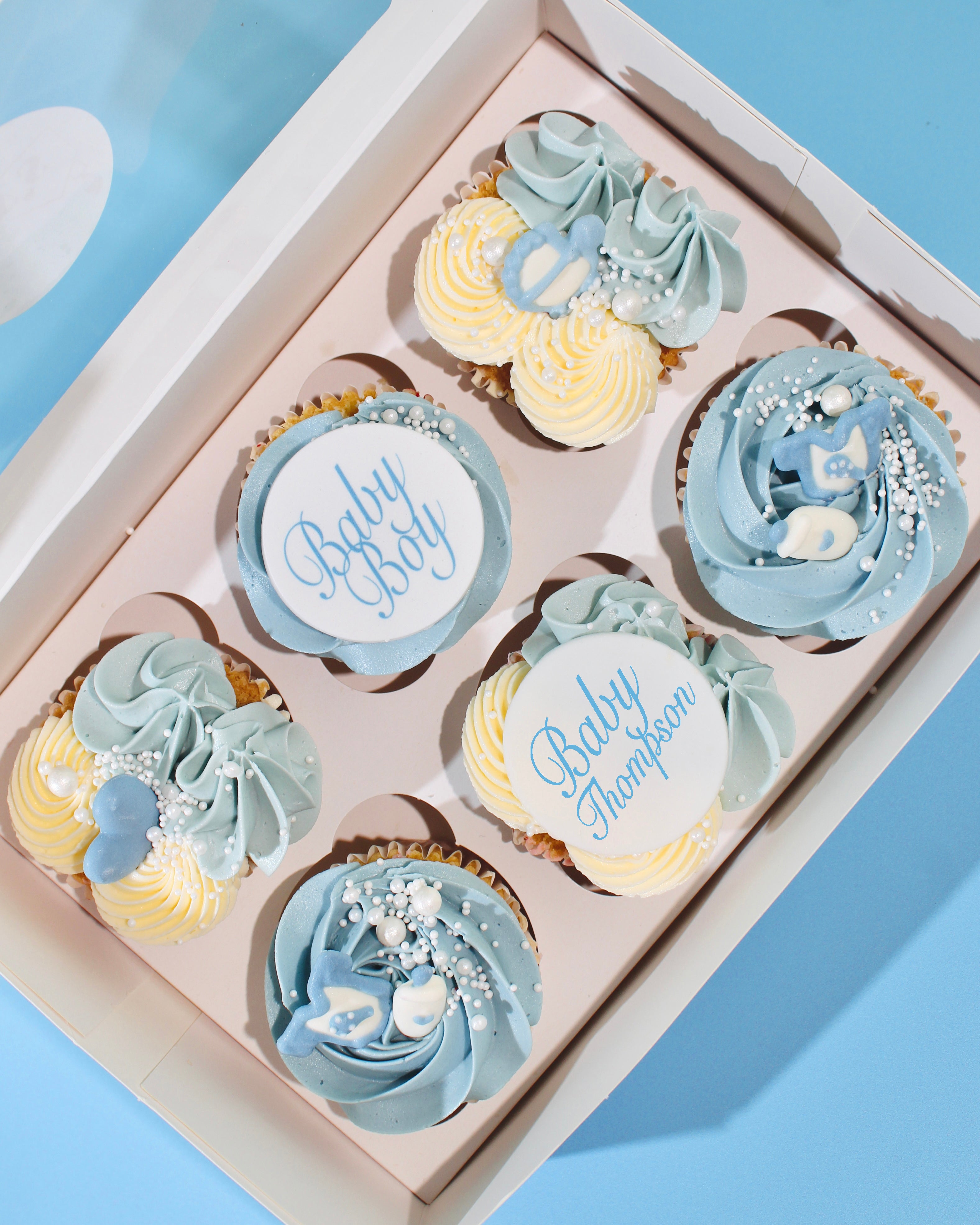 Baby Boy cupcakes with blue and cream buttercream and baby-themed decorations in gift box.