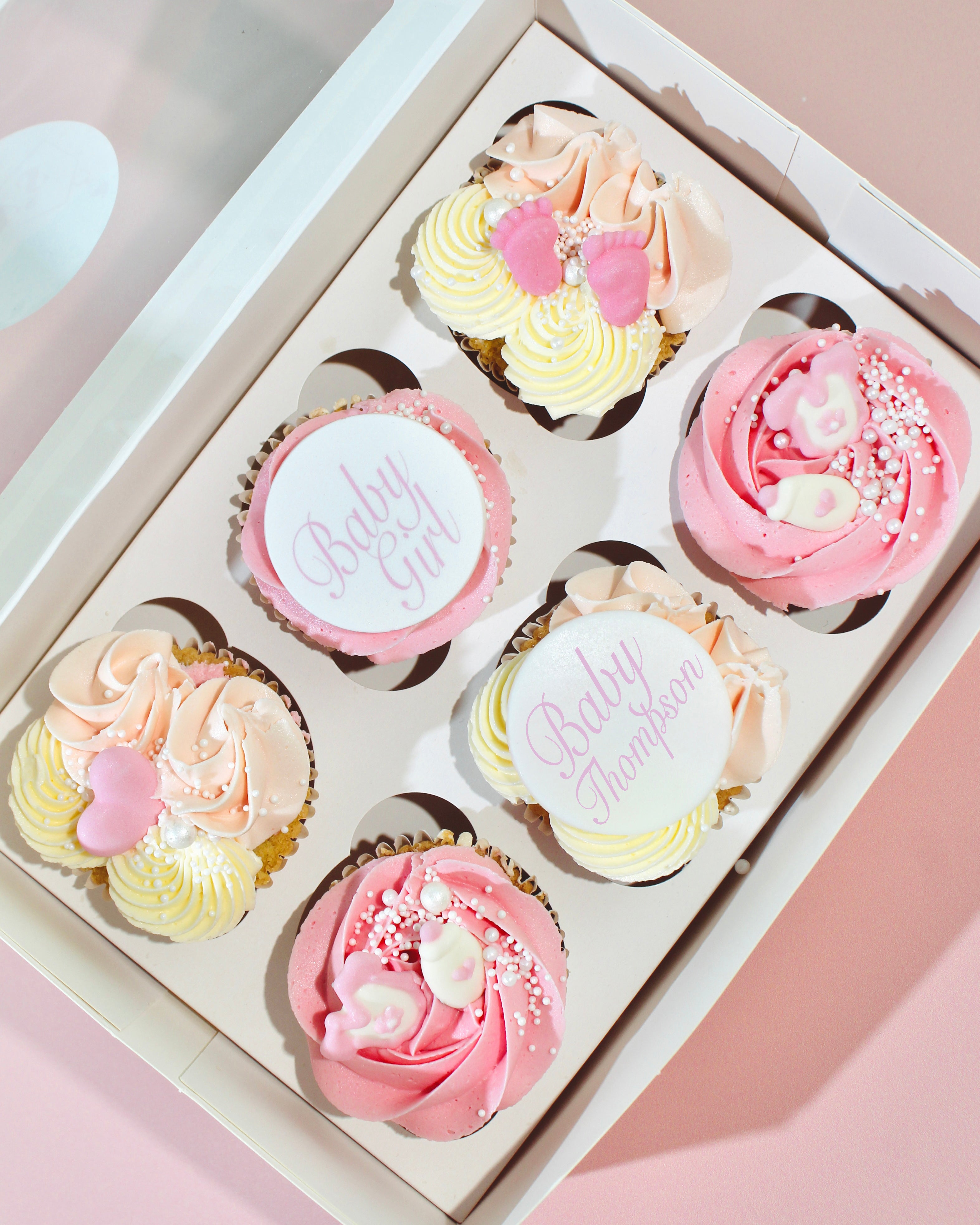 Box of pink Baby Girl Cupcakes decorated with buttercream swirls, sugar pearls and baby-themed toppers, including an optional personalised name topper.