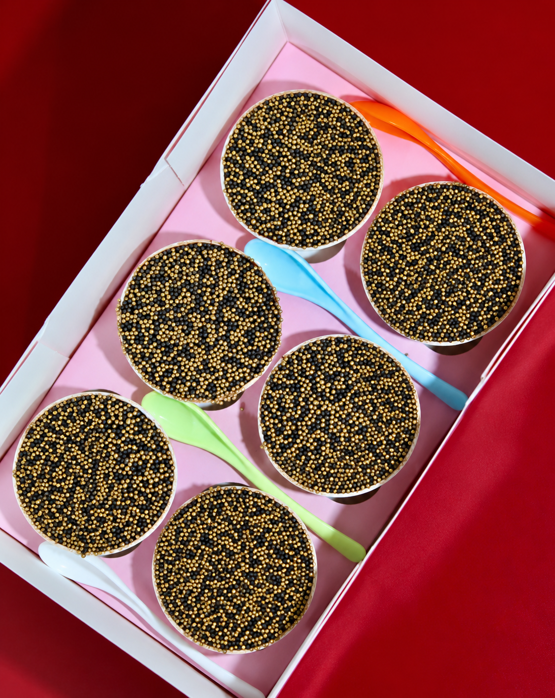 Six Bonfire Night Dot Cakes covered in black and gold sprinkles in open white gift box with colourful spoons