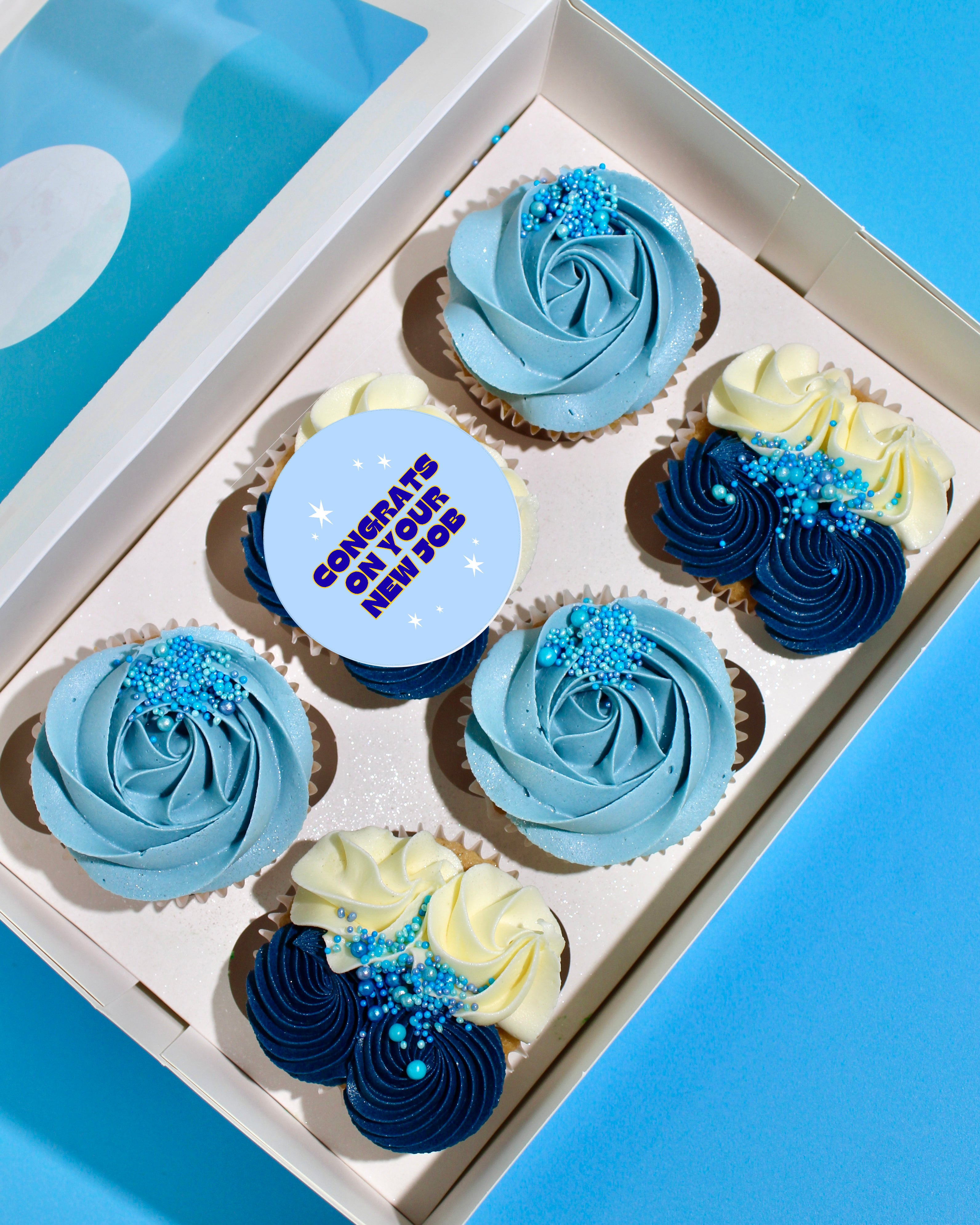Box of blue New Job cupcakes with buttercream swirls and “Congrats on Your New Job” edible topper.