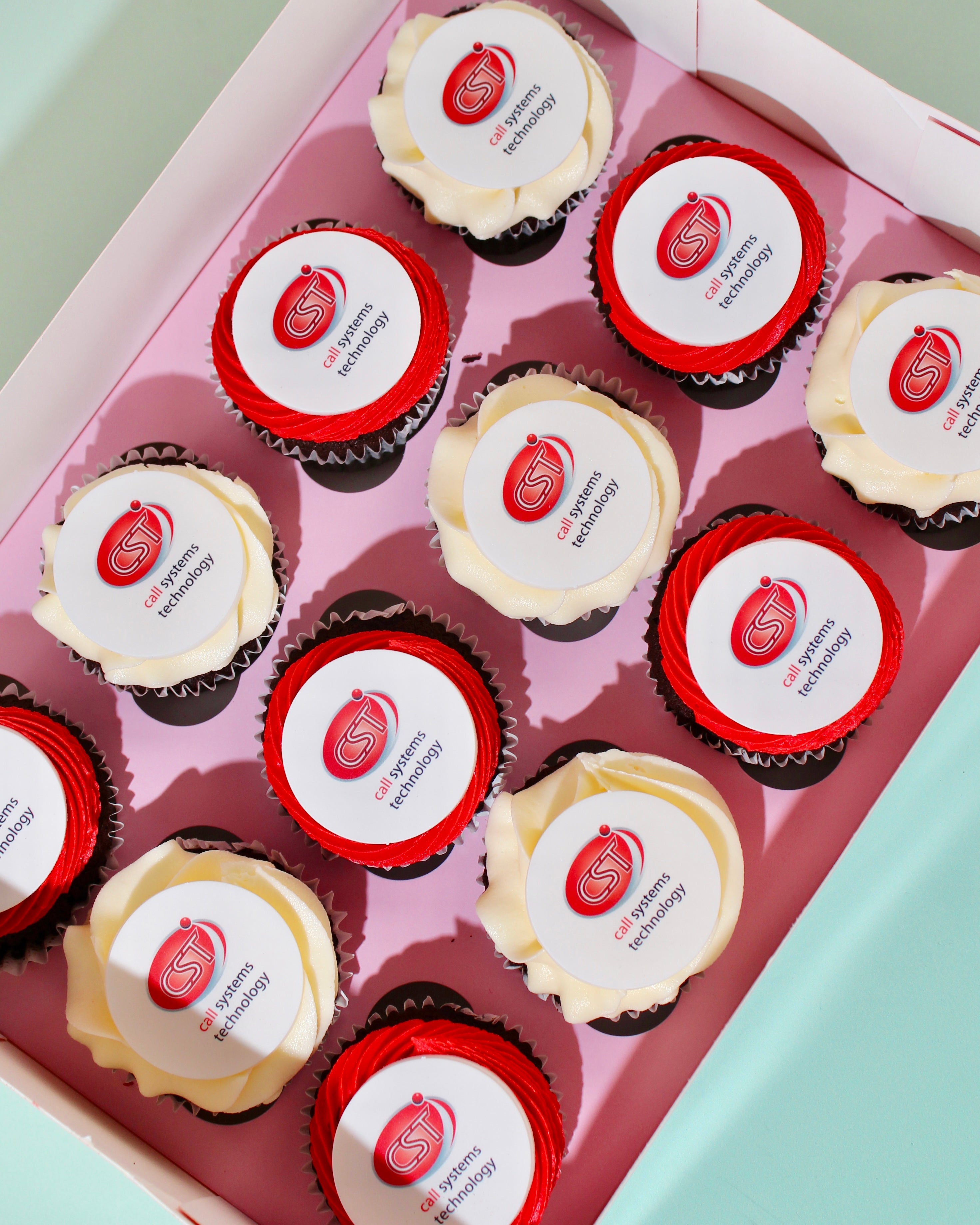 A box of cupcakes arranged in a pink tray, each topped with white frosting and a red circular logo. 