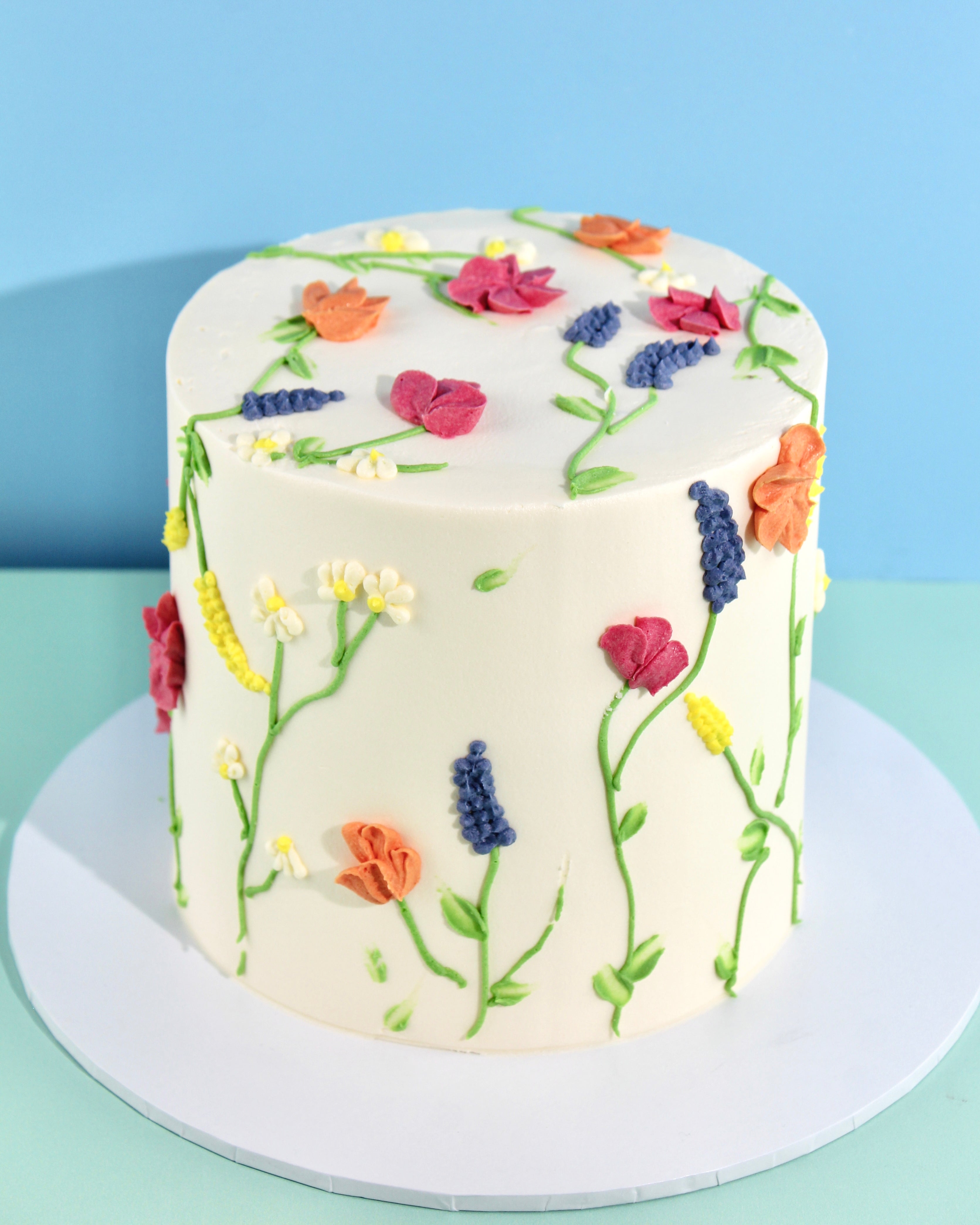 White buttercream Wildflowers Cake decorated with hand-piped colourful blooms and green stems, featuring delicate floral designs around the sides and top.