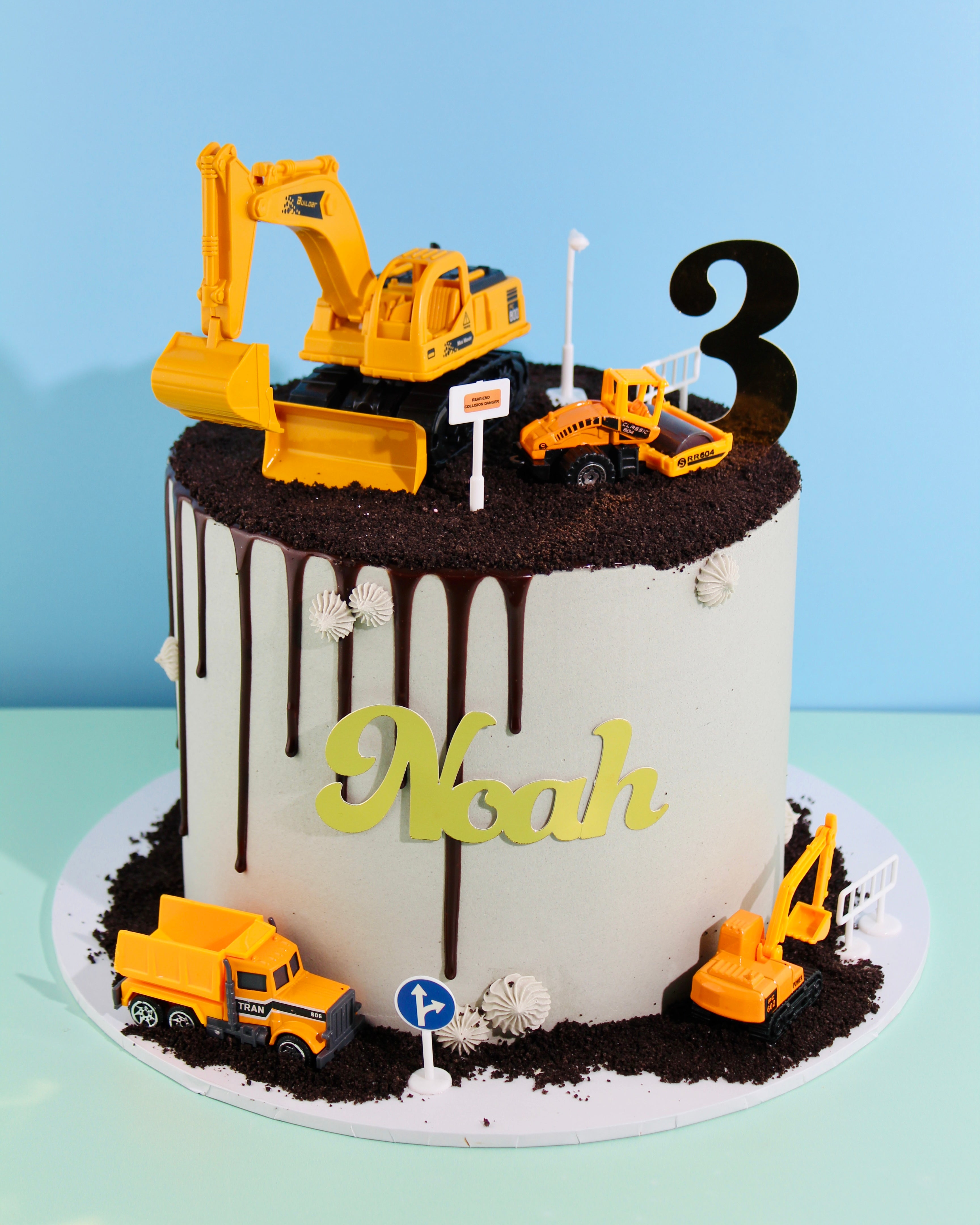 Construction-themed birthday cake with digger and truck toppers, chocolate dirt, personalised name, and number decoration