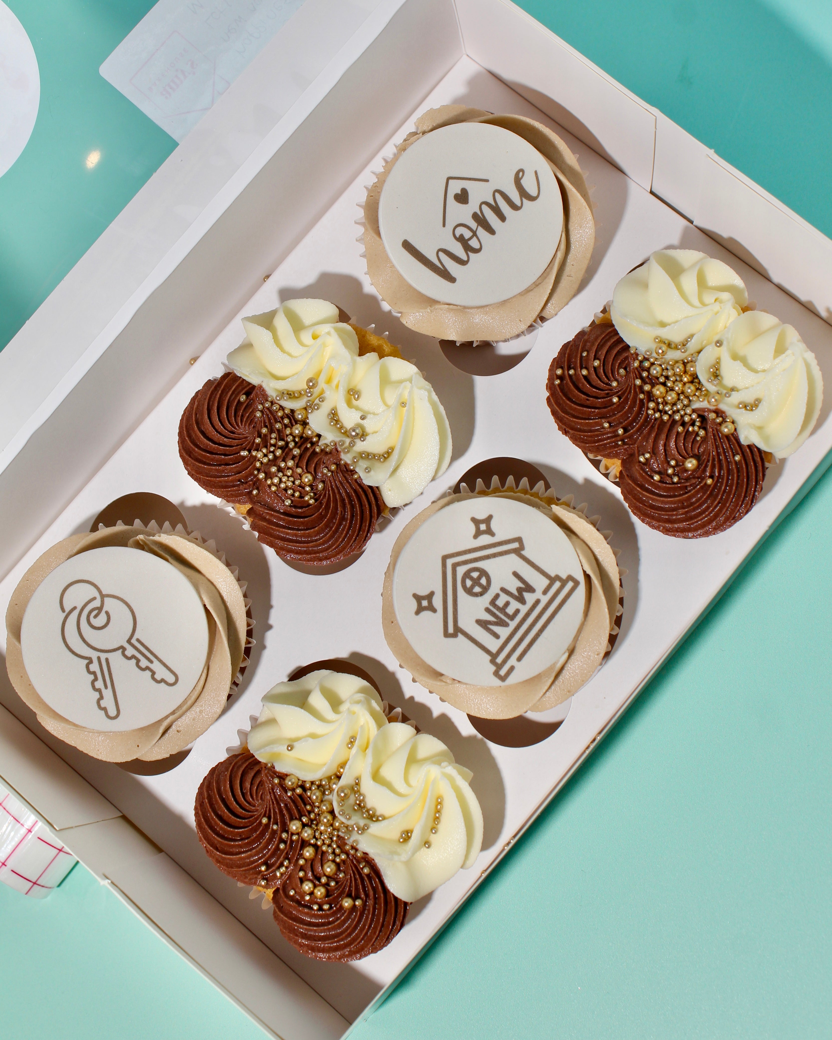 Box of six New Home cupcakes with “Home”, “Keys” and “New Home” edible toppers in beige, chocolate and vanilla icing.
