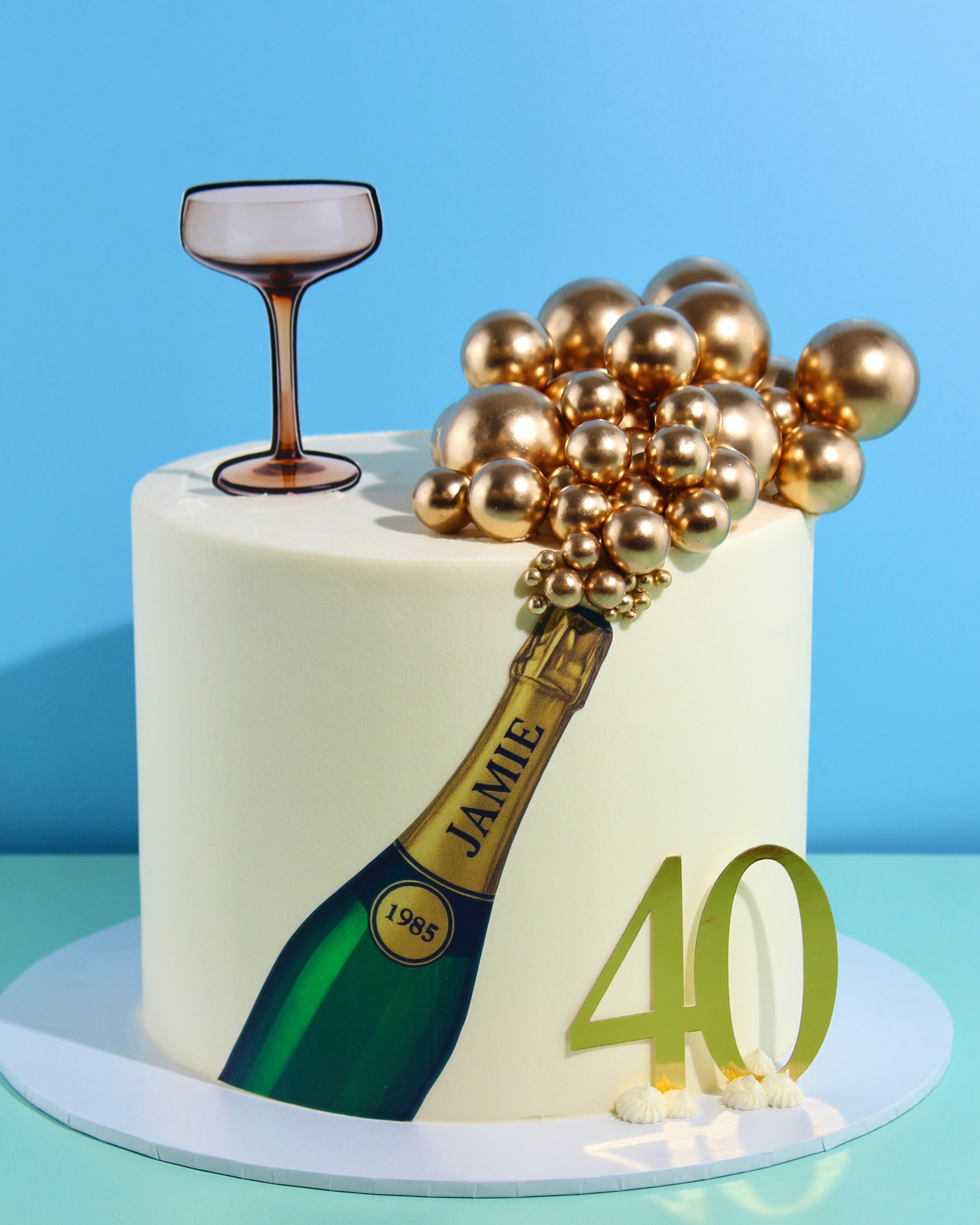 Champagne-themed birthday cake with gold spheres, personalised champagne bottle design, coupe-glass topper, and gold number decoration