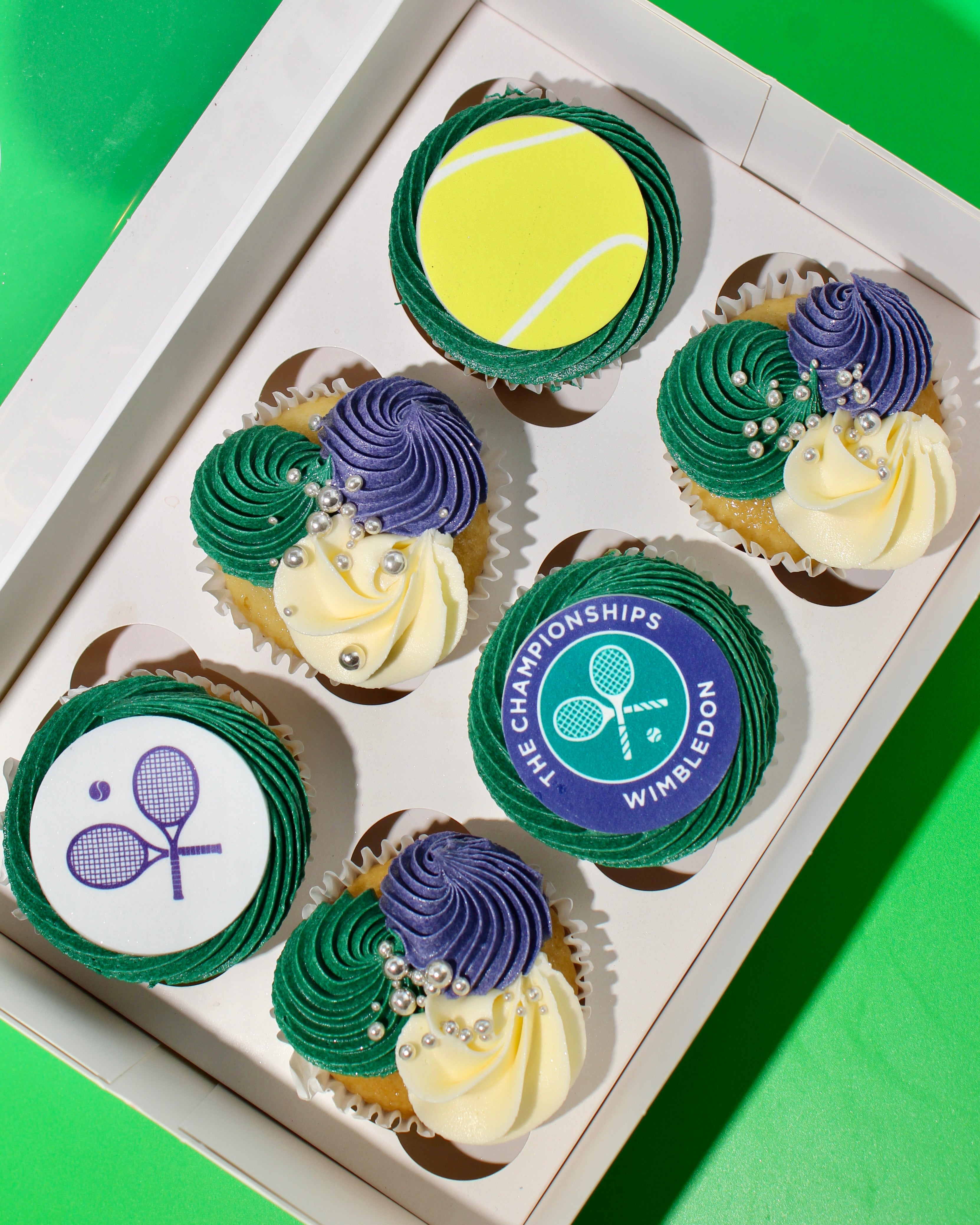 Six Wimbledon themed cupcakes with green and purple rosettes and tennis racket disc in open gift box
