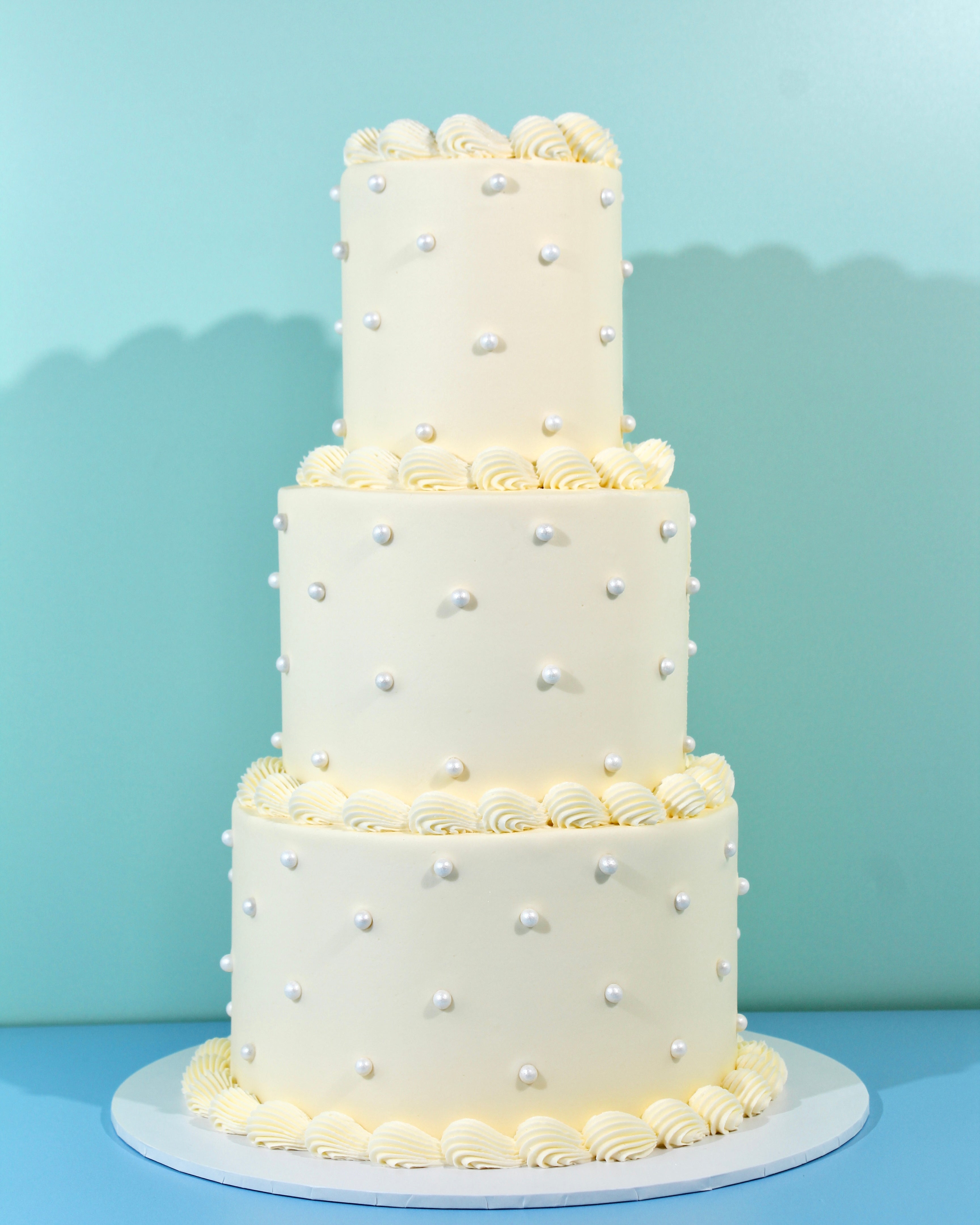 Three-tier ivory wedding cake decorated with pearl accents and piped buttercream borders on a white cake stand.