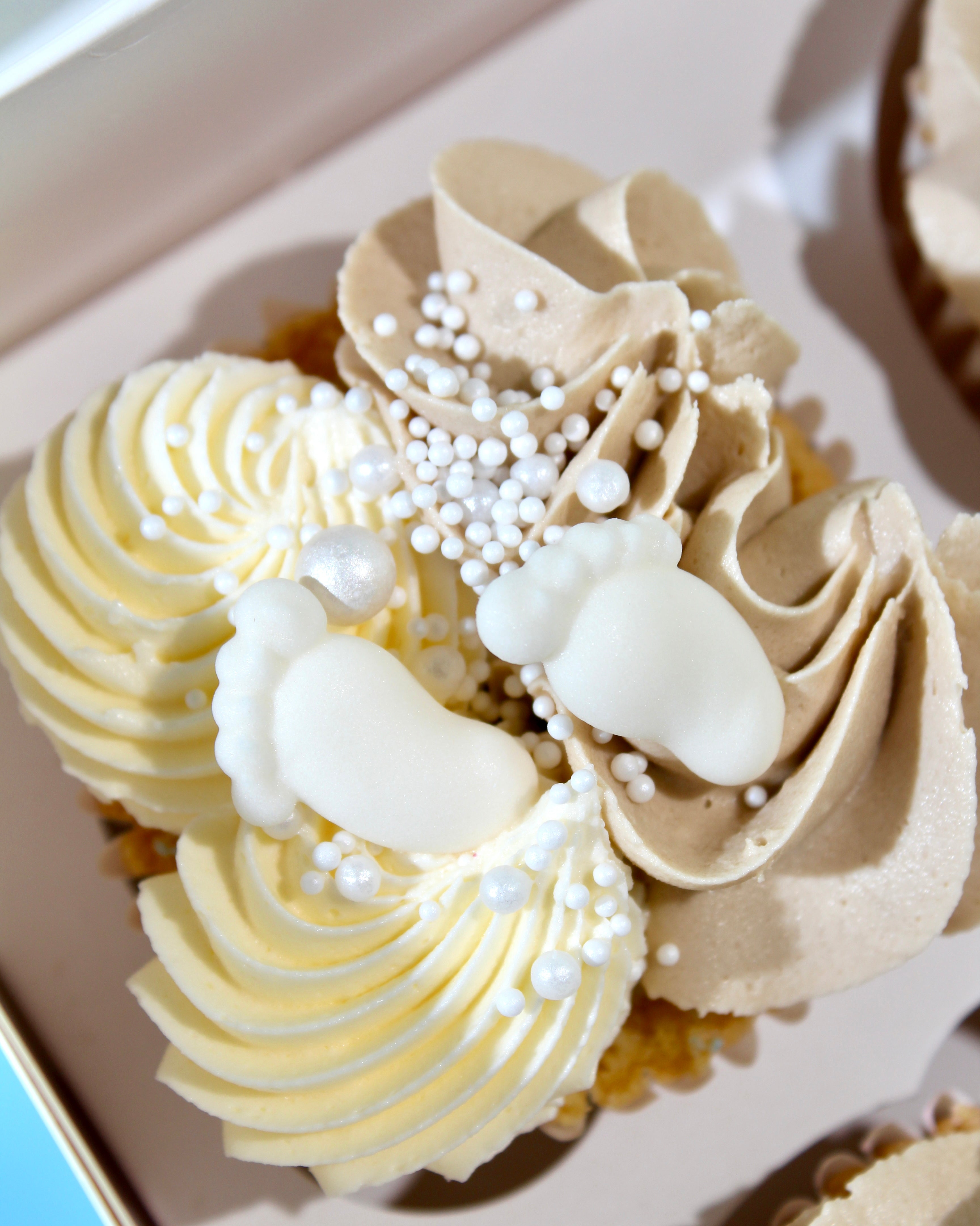 Box of baby shower cupcakes decorated in beige, cream and white with baby feet