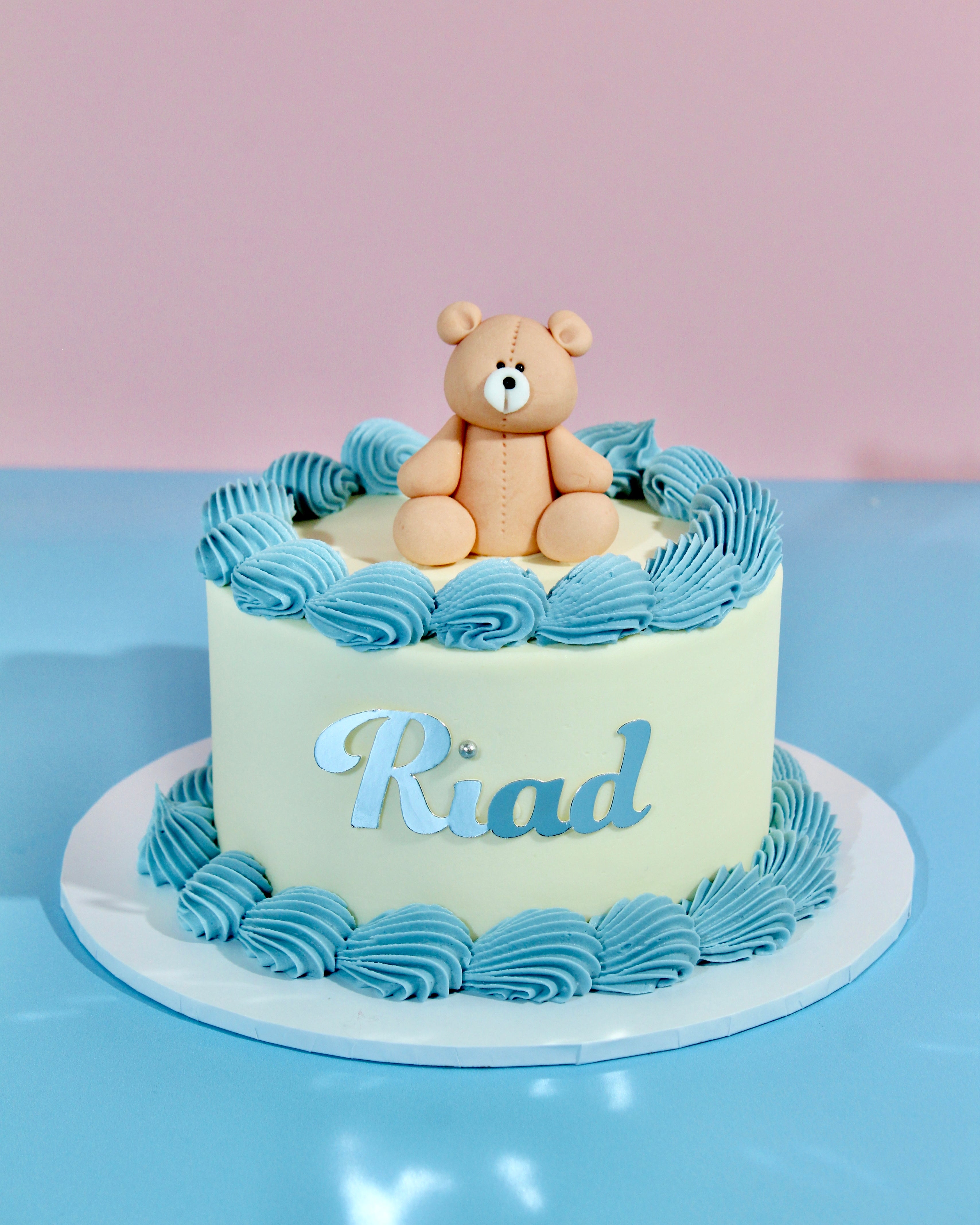 Cake with a teddy bear on top and the name 'Riad' written on it, against a light pink and blue background.