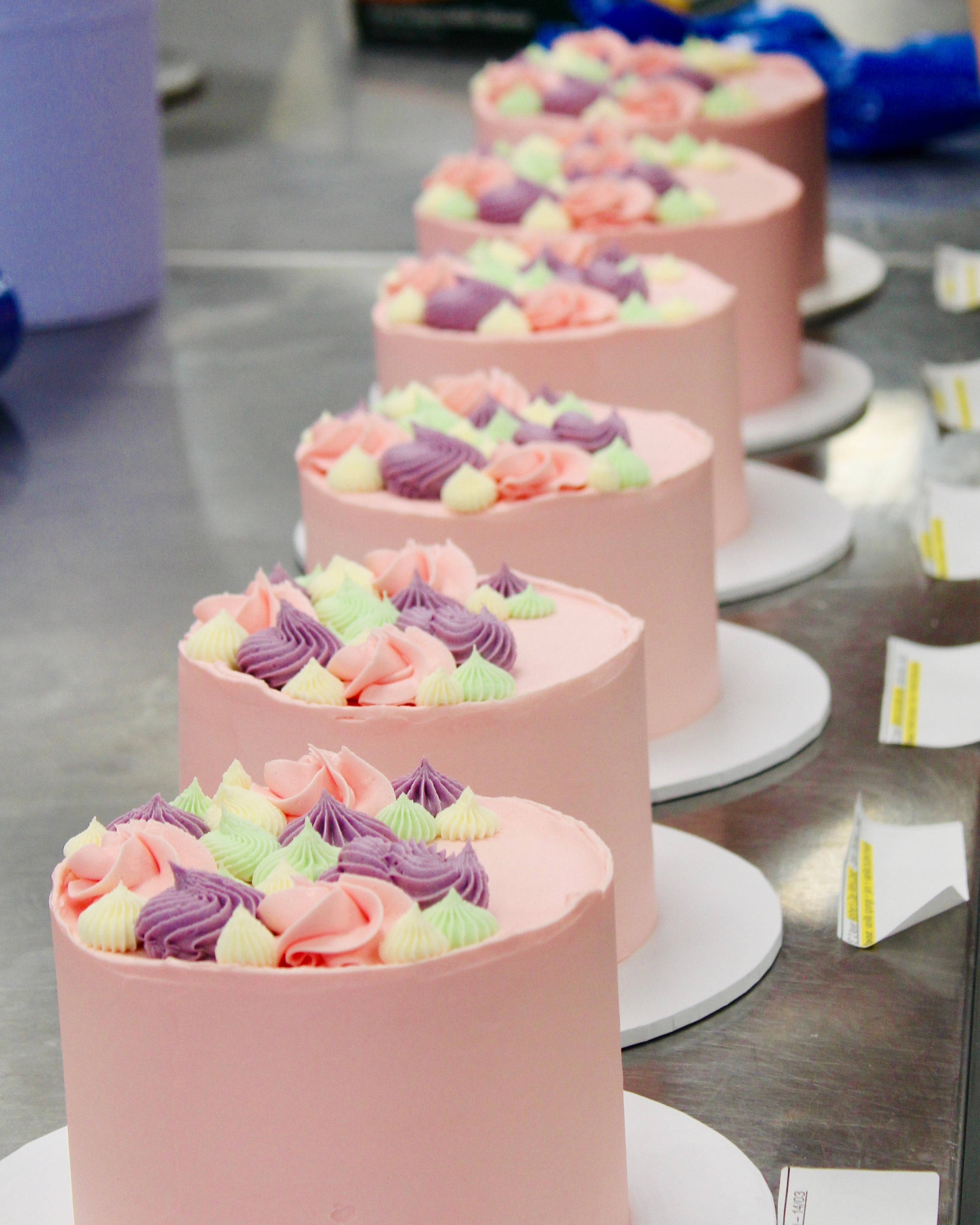Row of small pink cakes with colorful frosting on a metal surface.