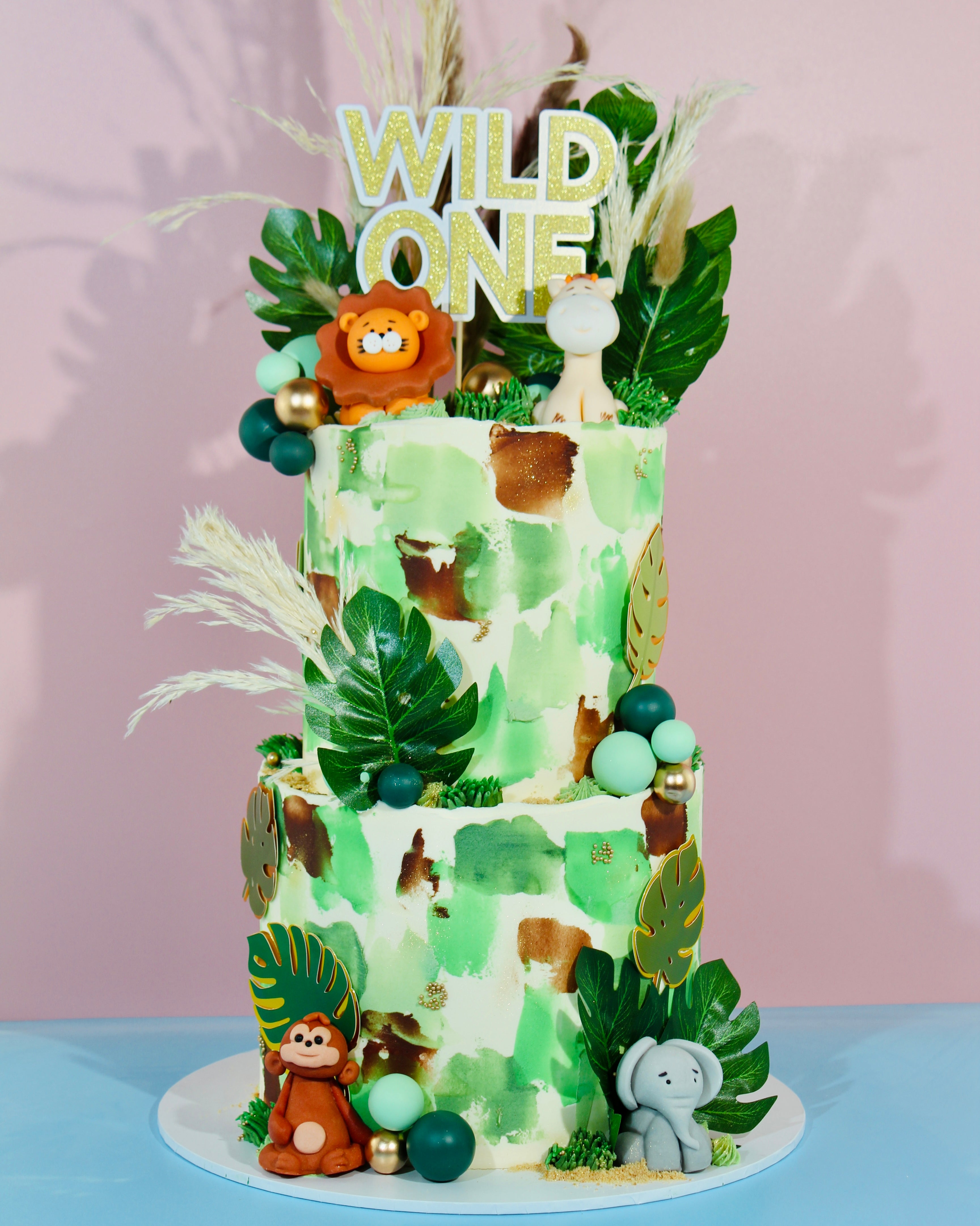 Two-tier jungle safari first birthday cake with Wild One topper, tropical leaves and animal figurines