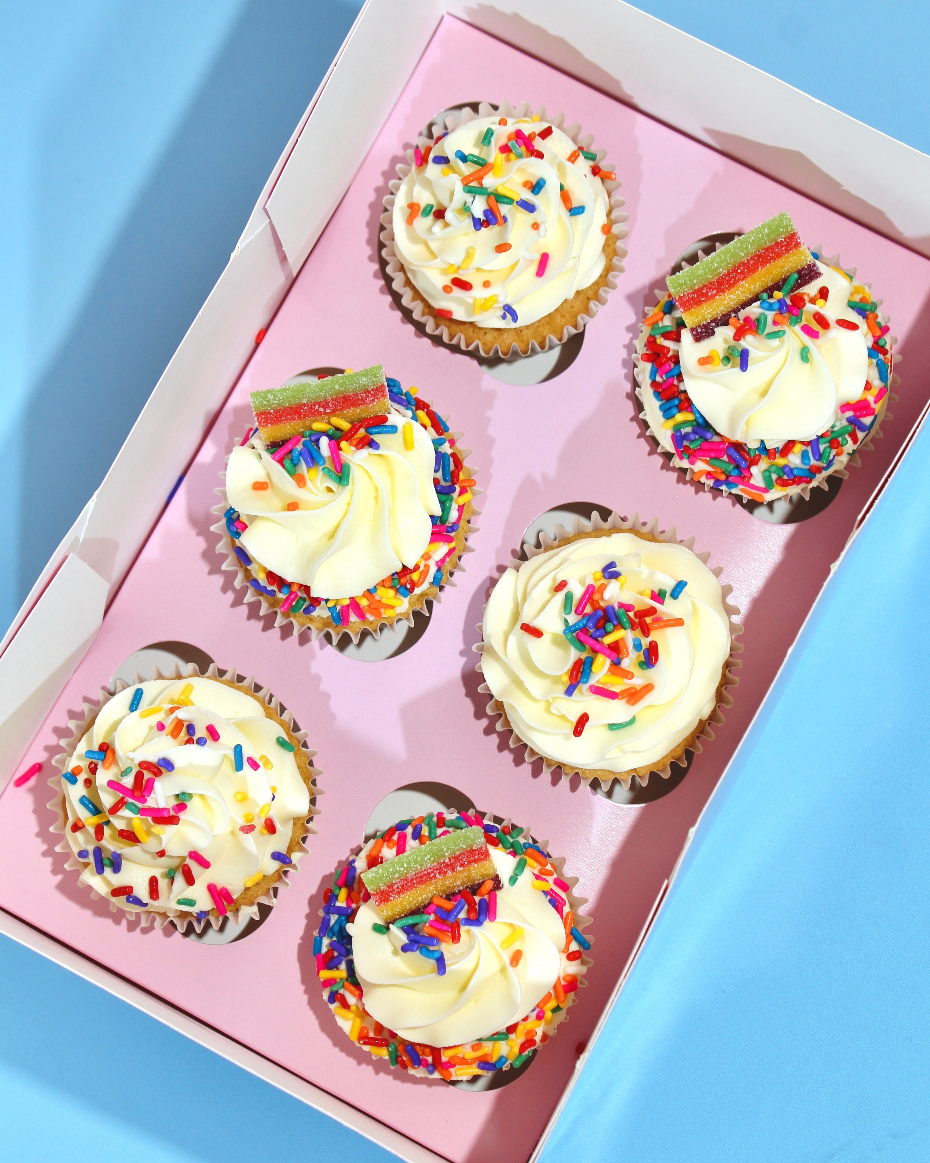 Six cupcakes with white frosting and colorful sprinkles in a pink box on a blue background