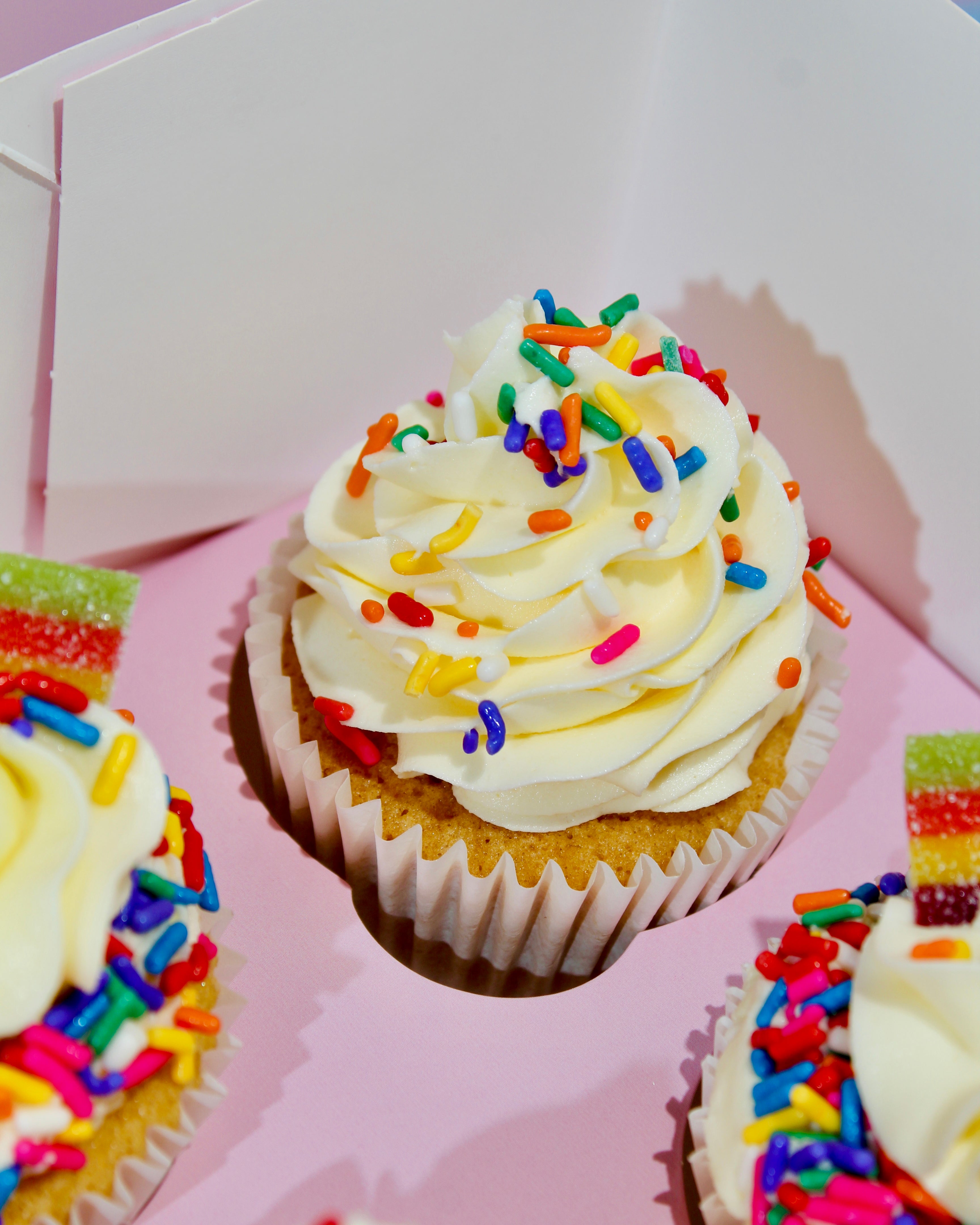 Cupcakes with white frosting and colorful sprinkles in a pink box.
