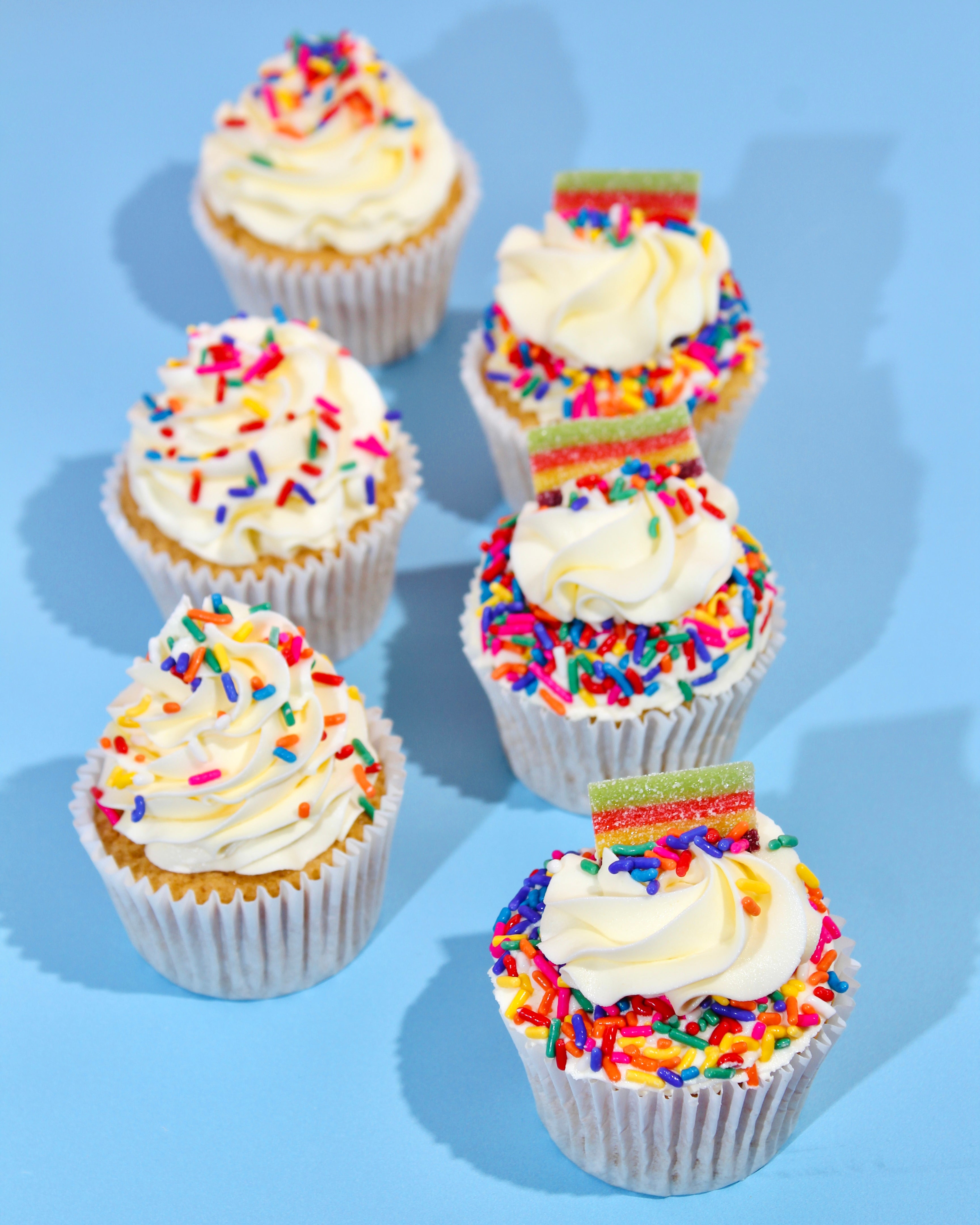 Six cupcakes with white frosting and colorful sprinkles on a blue background