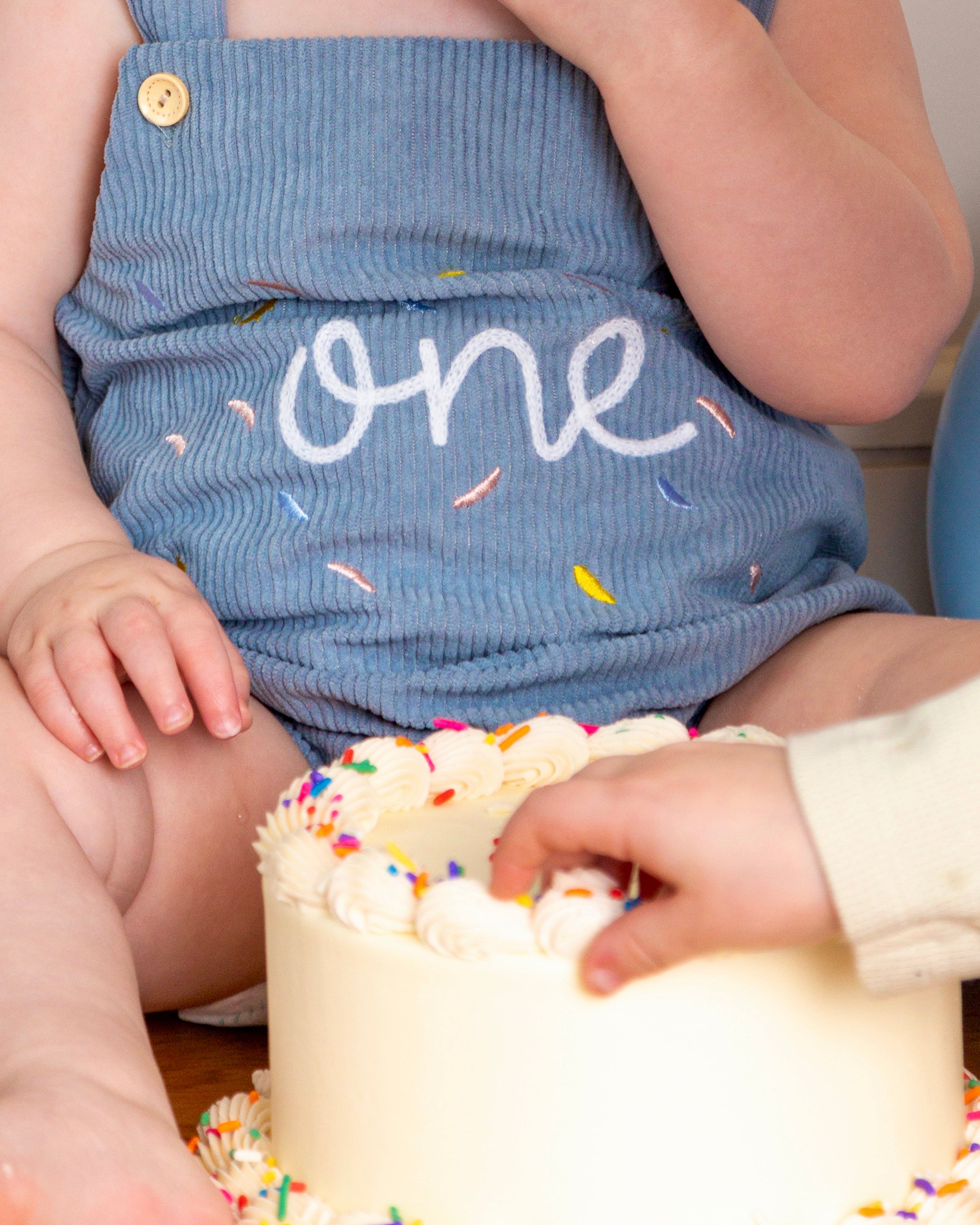 Child in a blue outfit with 'one' printed on it, reaching for a small birthday cake with colorful sprinkles.