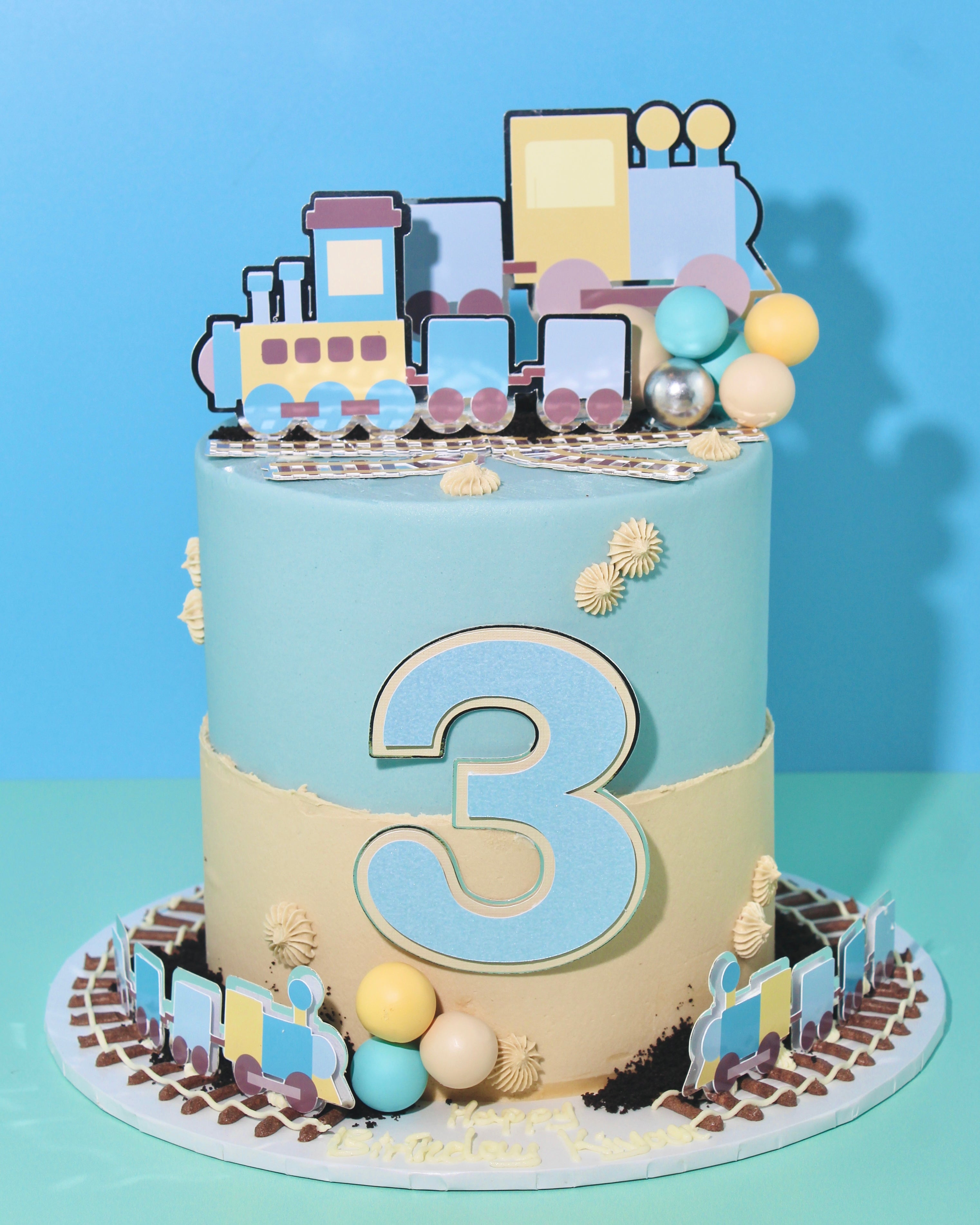 Pastel train-themed birthday cake with train toppers, buttercream tracks, piped details and a large number decoration