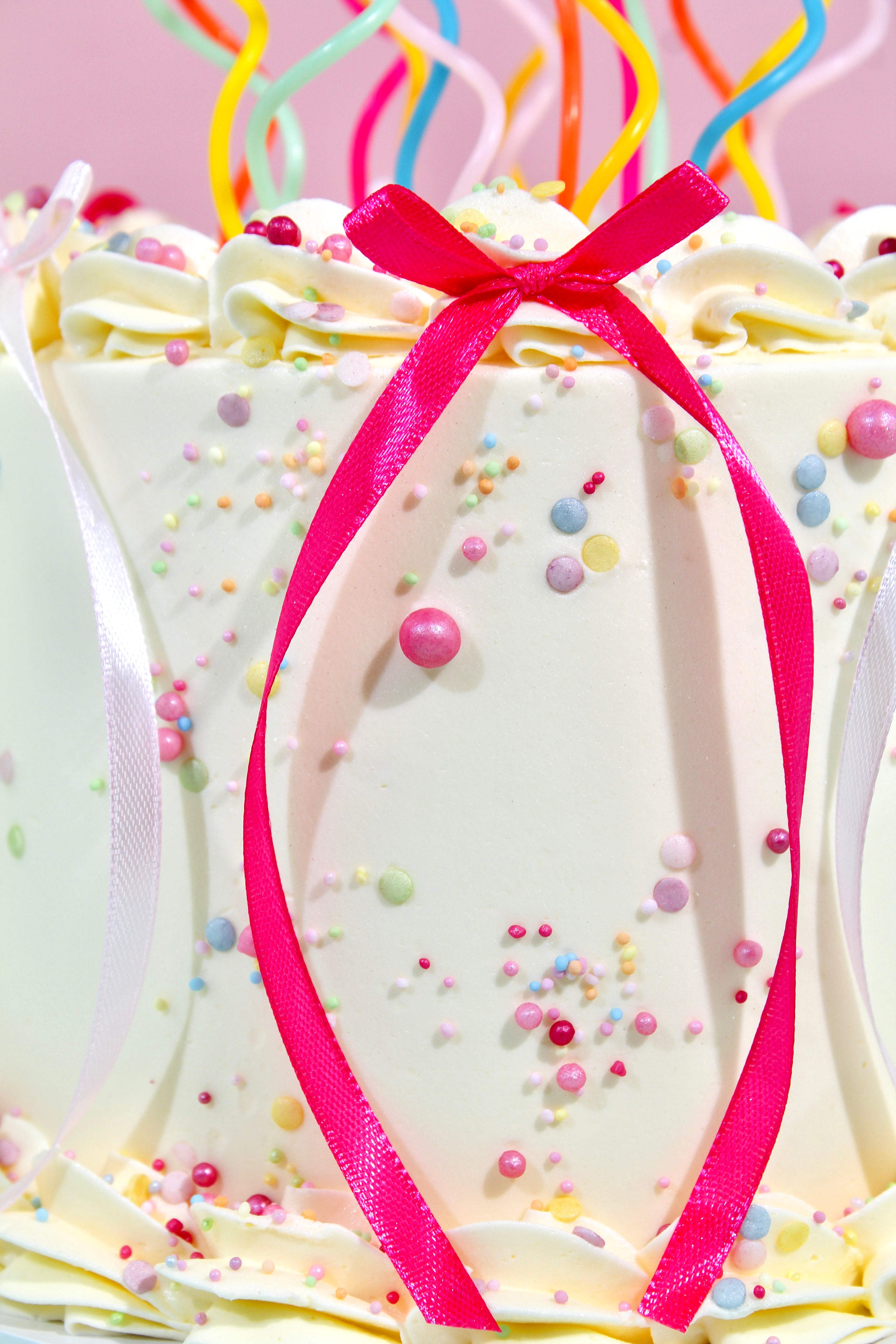 Decorative cake with white icing, pink ribbon, and colorful sprinkles on a pink background.