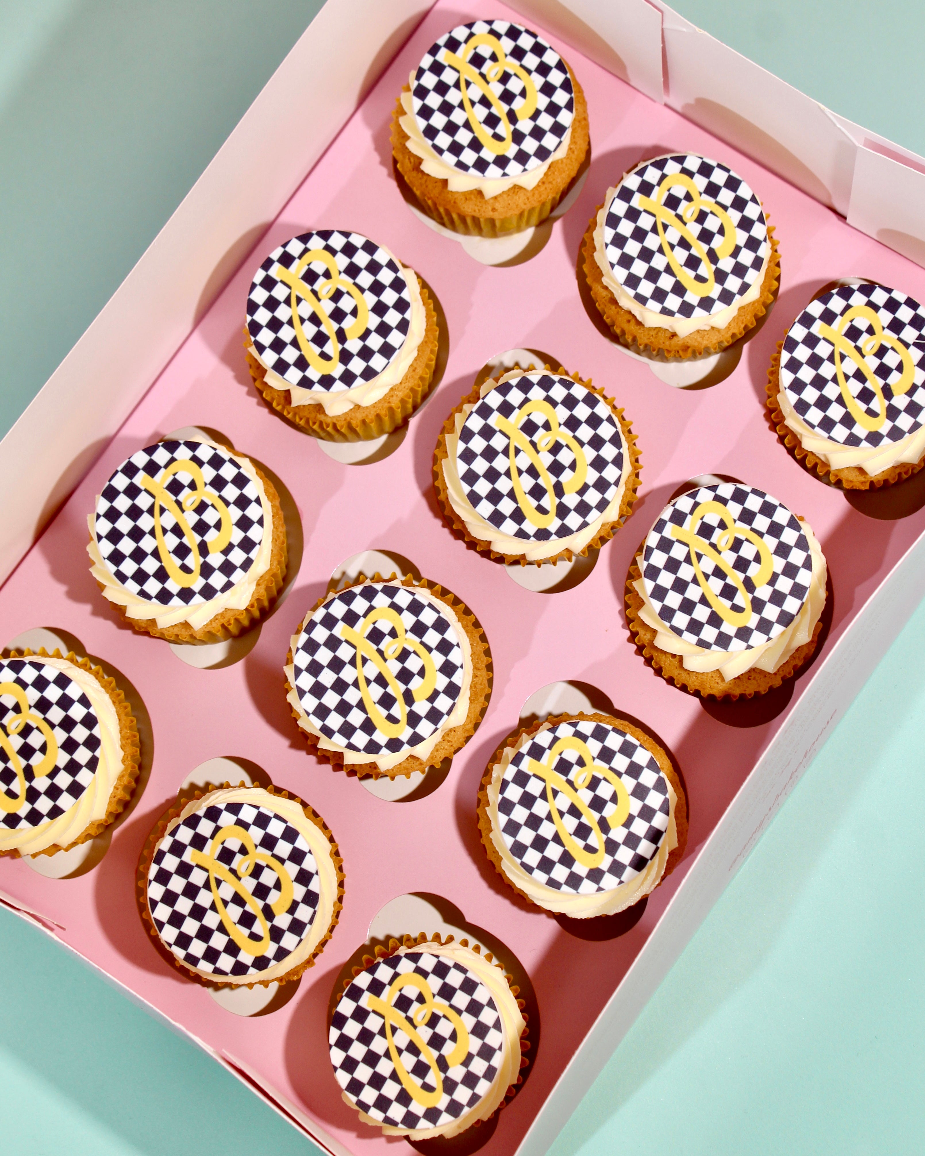 A box of cupcakes with cream frosting and black-and-white checkered toppers featuring a gold monogram, arranged in a pink tray.