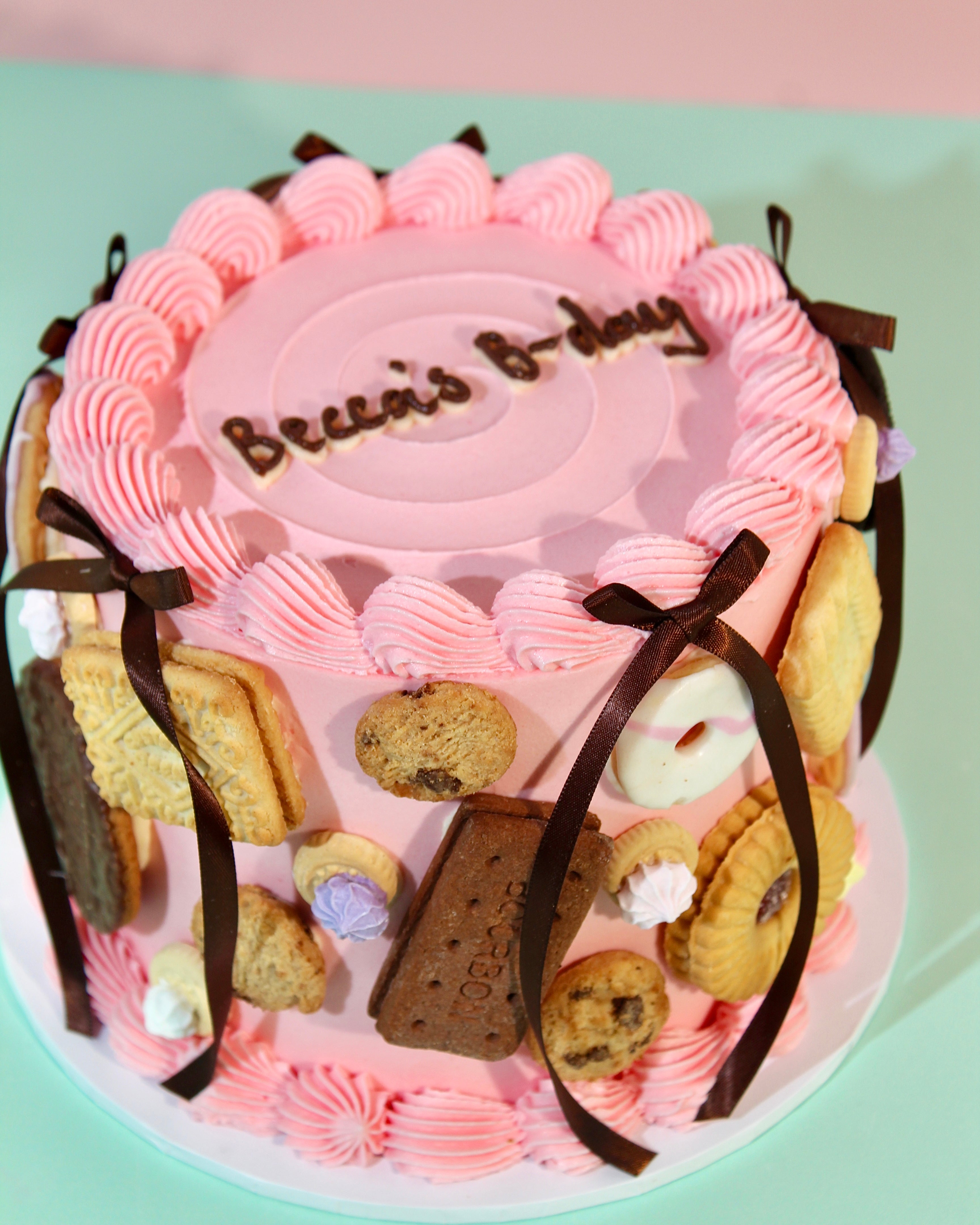Biscuit covered birthday cake with chocolate satin ribbon bows and pink piped buttercream borders from above