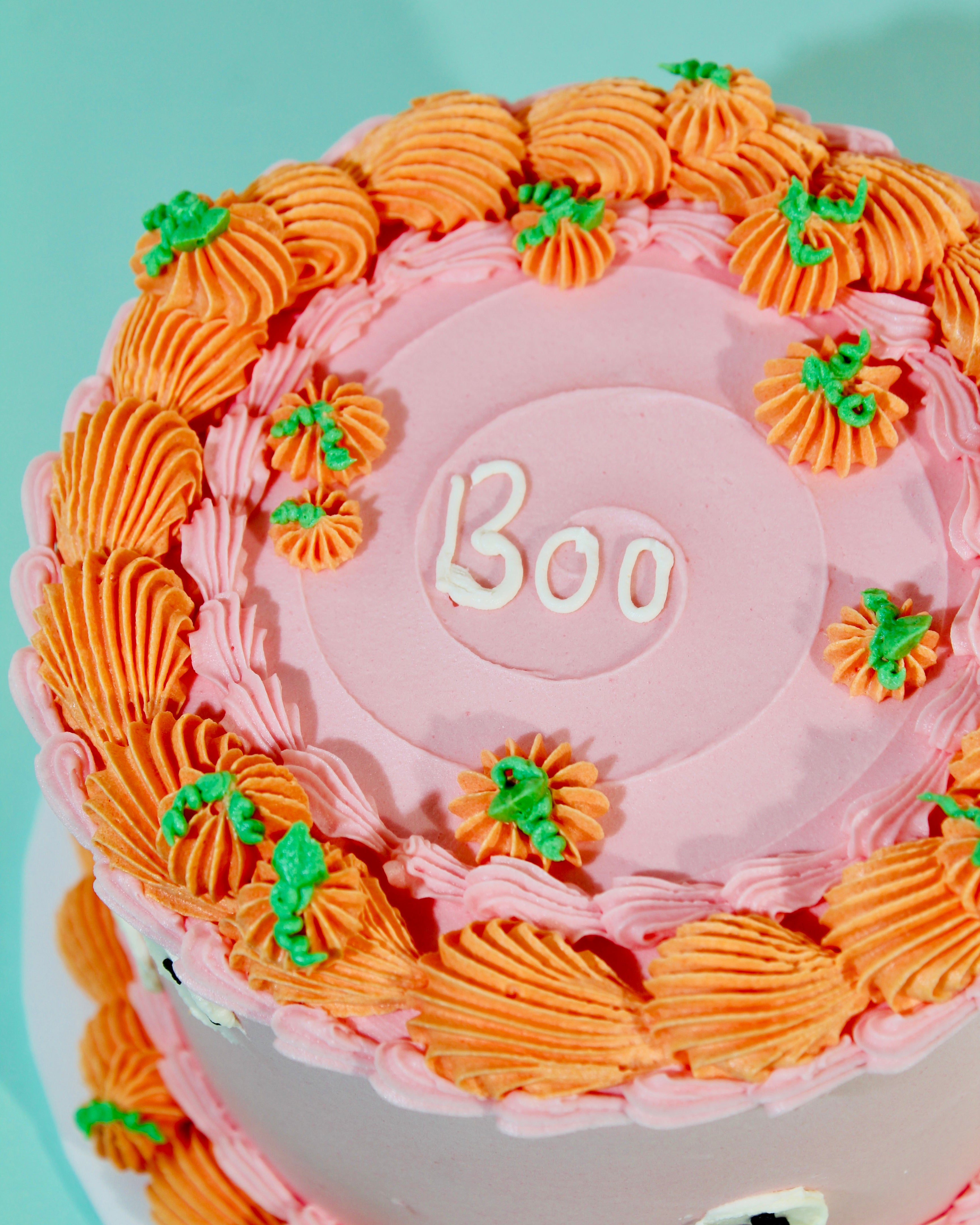 Close-up of Boo iced message on pink buttercream cake top surrounded by orange pumpkin rosettes with green leaves