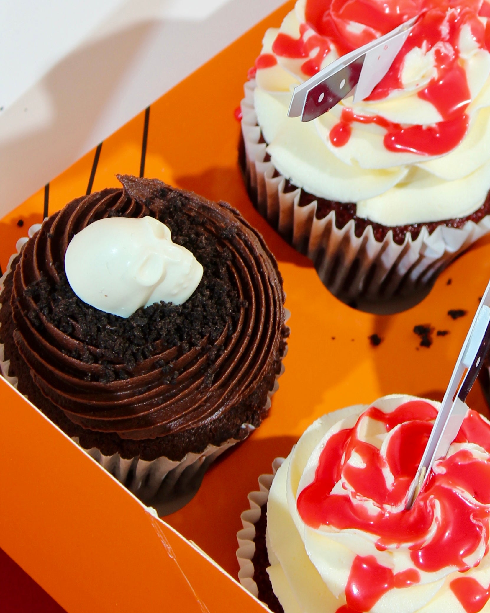 Close-up of chocolate skull cupcake alongside blood splatter white buttercream cupcake with mini axe charm in Halloween box