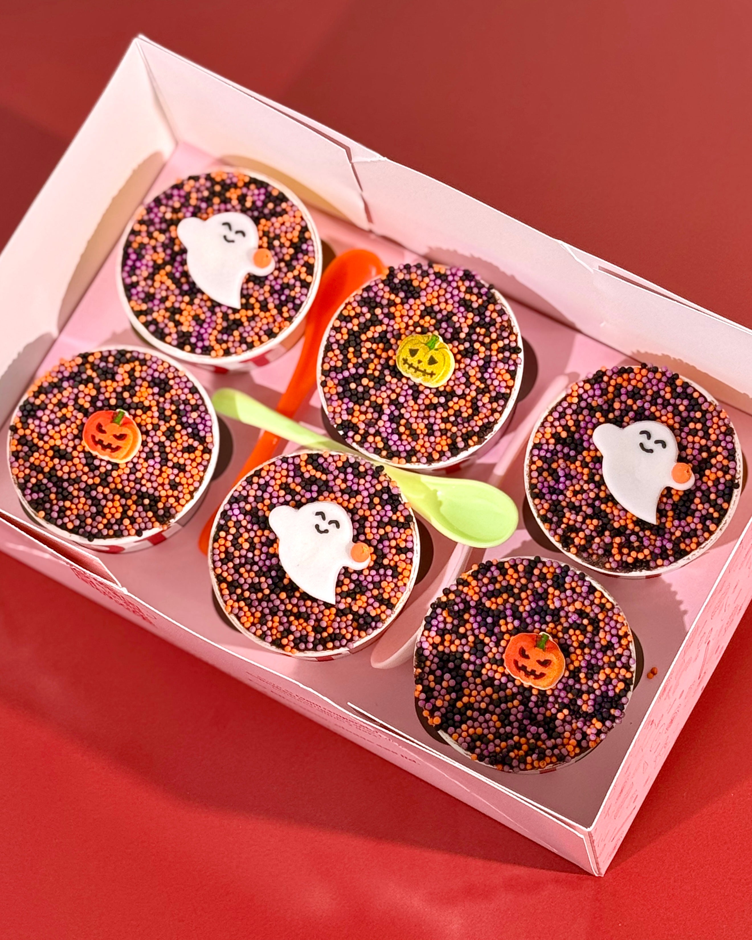 Six Halloween individual cake pots with ghost and jack-o-lantern toppers on Halloween dot sprinkle topping with colourful spoons