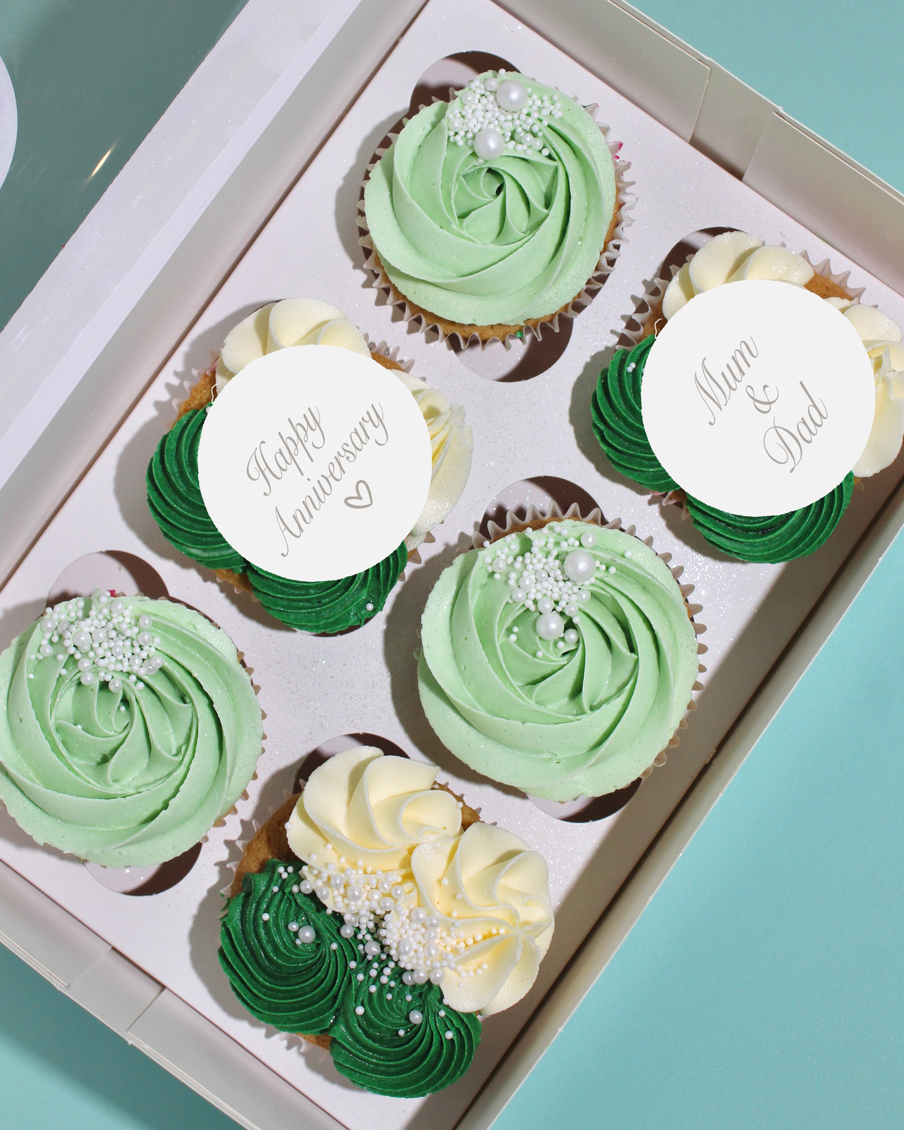 Box of anniversary cupcakes in green and cream tones with custom toppers and piped buttercream