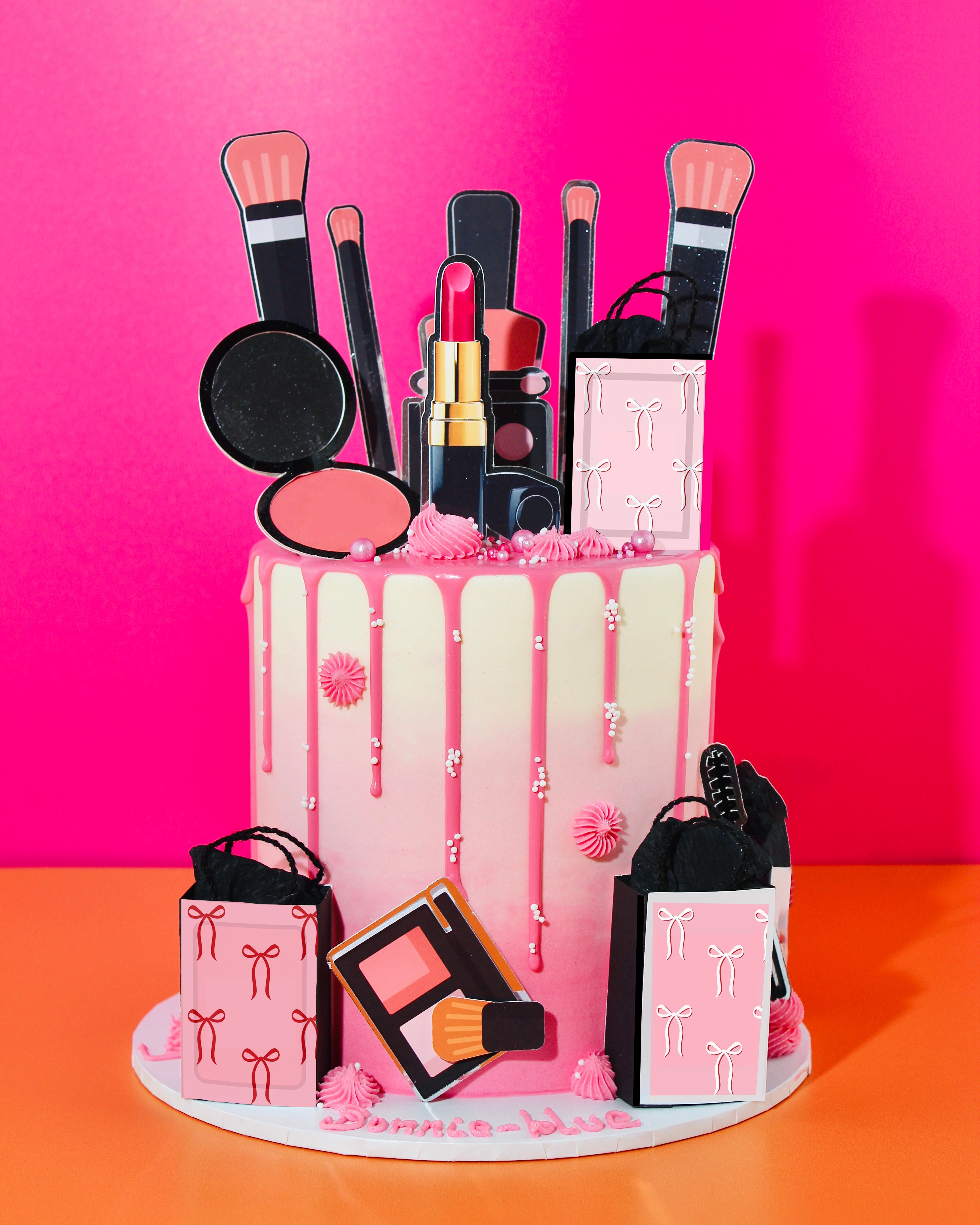 Pink drip make up cake decorated with beauty-themed toppers including lipstick, brushes, blush and mini gift bags.